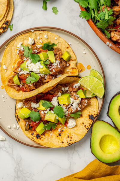 Two pulled pork tacos on a plate with queso fresco, cilantro, and avocado.
